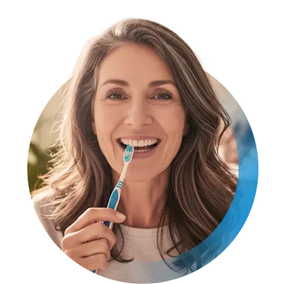 Frau mittleren Alters mit braunen langen Haaren lächelt und hat eine Zahnbürste in der Hand.