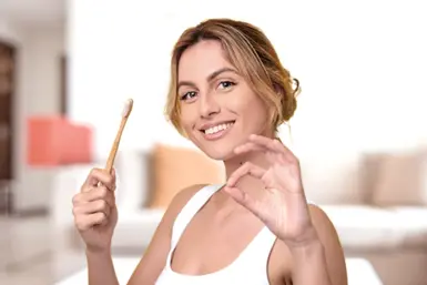 Junge Frau mit blonden Haaren und Dutt in einem weißten Top. Sie hält eine Zahnbürste aus Holz in der Hand und lächelt in die Kamera.