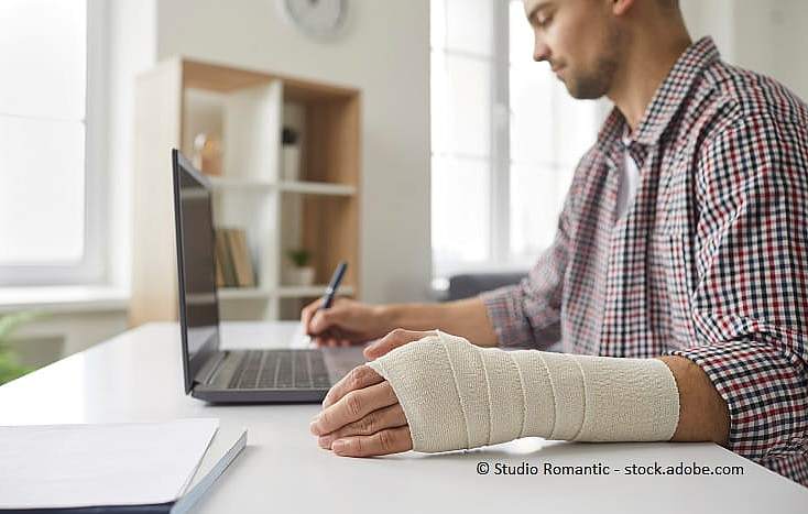 Bild eines Mannes mit gebrochenem Arm, der am Schreibtisch sitzt und etwas notiert.