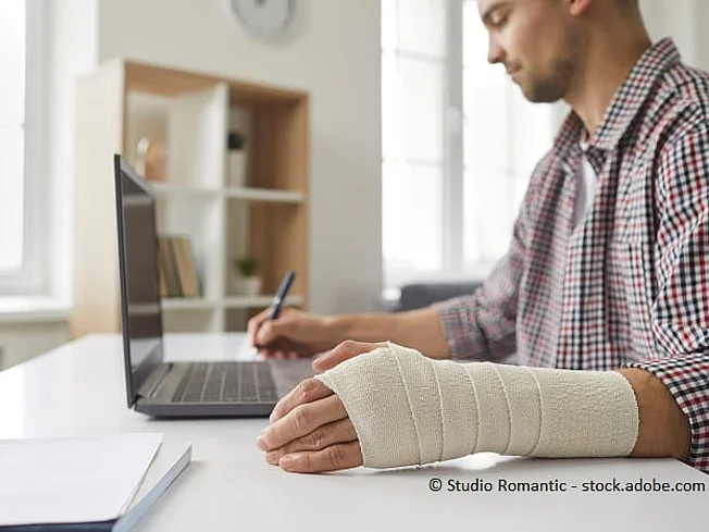 Bild eines Mannes mit gebrochenem Arm, der am Schreibtisch sitzt und etwas notiert.