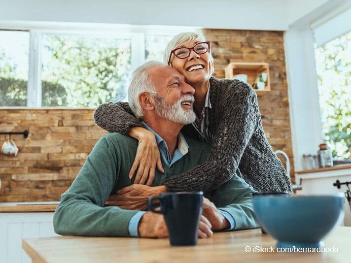 Ratgeber Pflege - Ältere Frau, die in ihrer Küche glücklich ihren Mann umarmt
