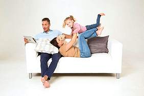 ZahnGesund - Münchener Verein  - Junge Familie mit eine kleinem Kind sitzt auf einem Ledersofa.
