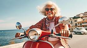 &Auml;ltere Dame mit Sonnenbrille sitzt lachend auf einem roten Motorroller.