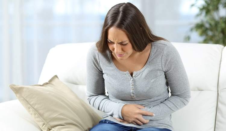 Bild einer Frau, die auf der Couch sitzt und ihren schmerzenden Bauch hält.