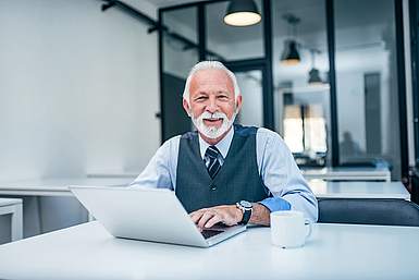 &Auml;lterer Herr mit grauen Haaren in Hemd und Weste sitzt am Laptop und lacht in die Kamera.