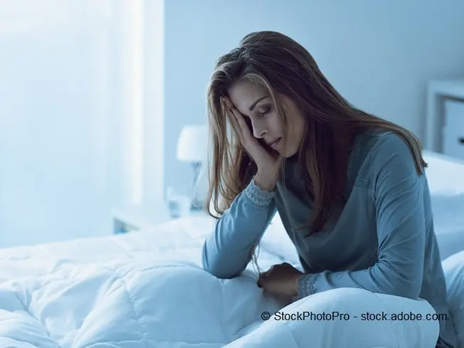 Bild einer Frau, die auf dem Bett sitzt und ihren Kopf auf ihren Arm stützt.