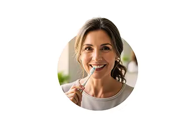 Frau mittleren Alters mit braunen lockigen Haaren und einem Zopf lächelt in die Kamera und hat eine Zahnbürste in der Hand.