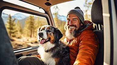 Mann mit Jacke und M&uuml;tze h&auml;lt seinen Hund und lacht in die Kamera.