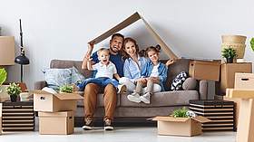 Junge Familie mit zwei Kindern sitzt auf der Couch unter einem Kartondach und lacht in die Kamera.