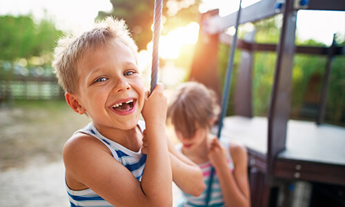 Kleiner Junge mit Zahnlücke beim Schaukeln auf dem Spielplatz - Zahnersatz für Kinder bei Unfällen