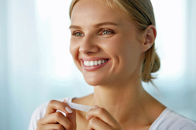 Schöne lächelnde Frau mit gesunden weißen Zähnen beim Auftragen des Zahnweißstreifens. 