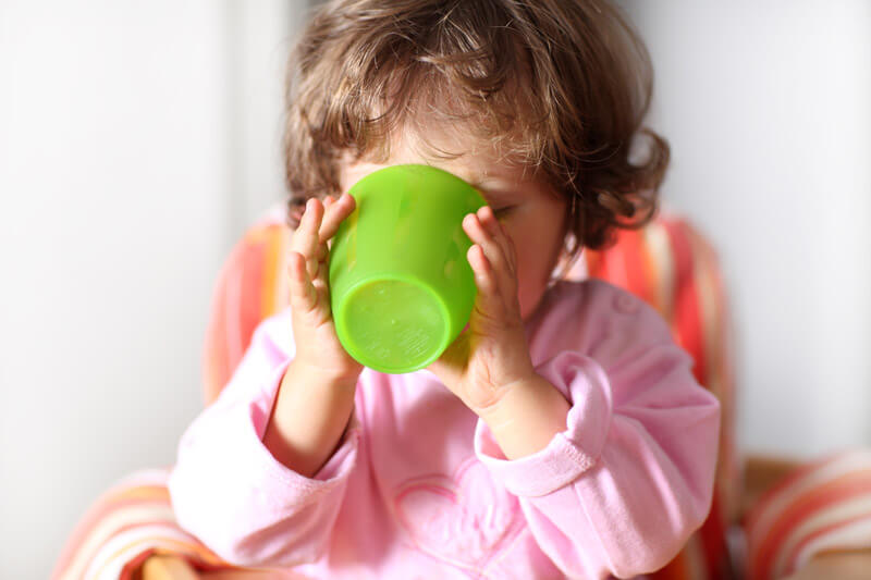 Zahnpflege Babys_Kleinkind im Kinderstuhl trinkt eigenständig aus einem grünen Trinkbecher.
